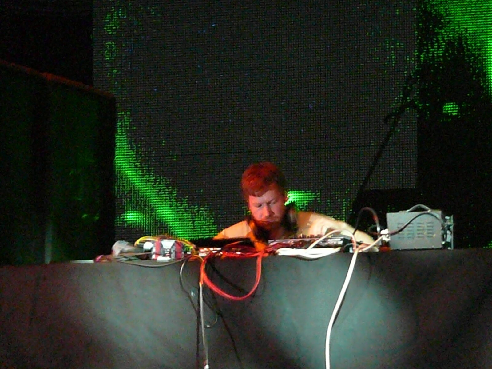 Aphex Twin performing behind a table of equipment in front of a green video screen