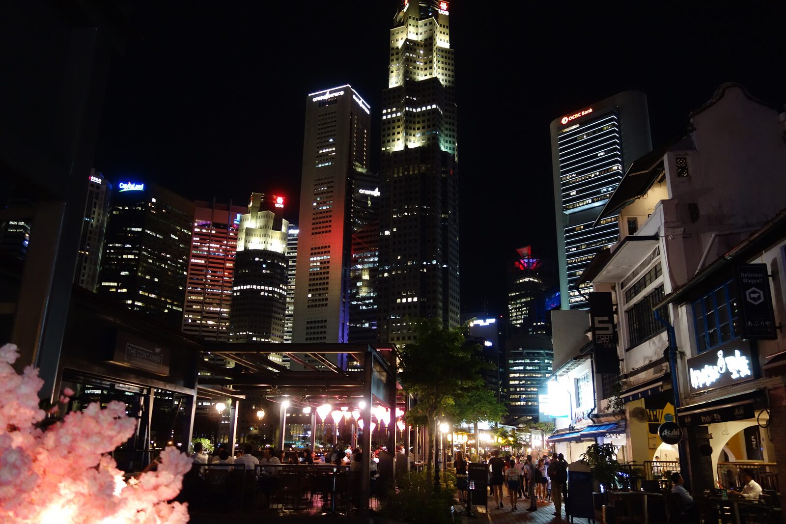Night view of the Boat Quay waterfront in Singapore, with lit shophouse bars and restaurants in the foreground and central business district office towers rising behind them.