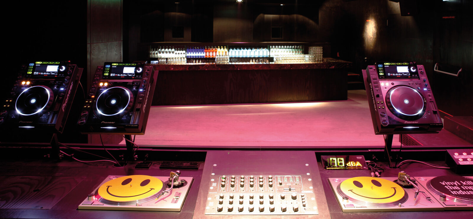 An empty DJ booth in a dark Munich techno club, with two CDJ players on each side, two turntables with yellow smiley slipmats, a rotary mixer in the middle and a lit bar in the background