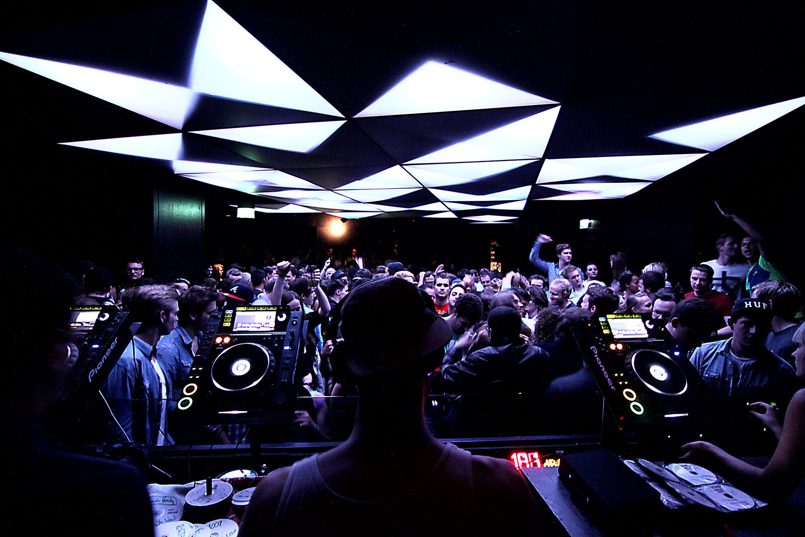 A crowded dance floor seen from behind a DJ in a Munich techno club, with three CD players in the foreground and angular white ceiling panels above the crowd