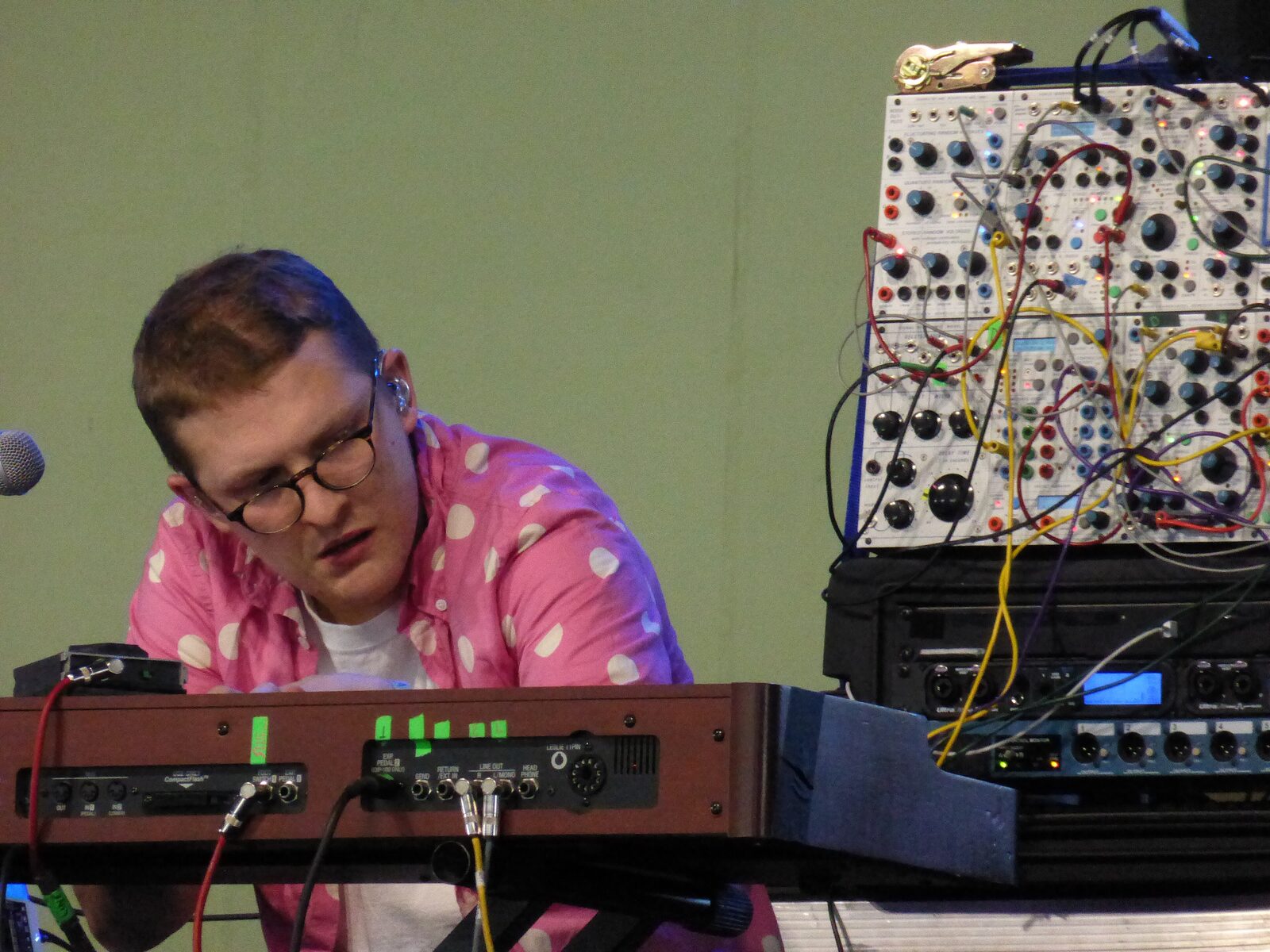 Floating Points in a pink polka-dot shirt and round glasses, leaning over a keyboard with a modular synthesizer rack beside him