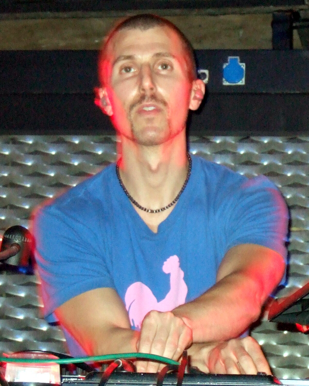 Josh Wink in a blue t-shirt with a pink rooster print, leaning over DJ equipment under red light in 2007