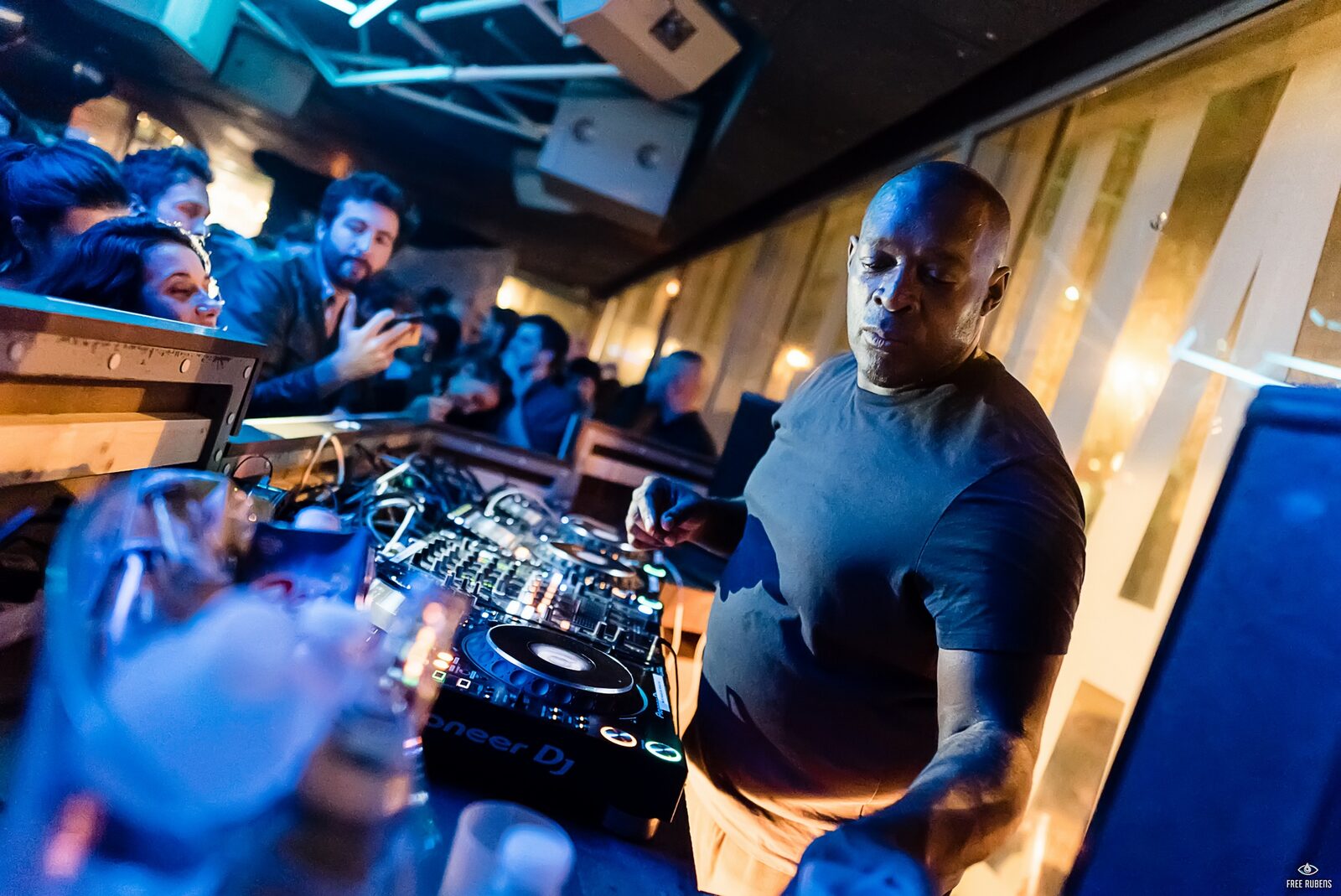 Kevin Saunderson in a dark t-shirt playing on Pioneer DJ equipment in a club, with people leaning in over the booth at left