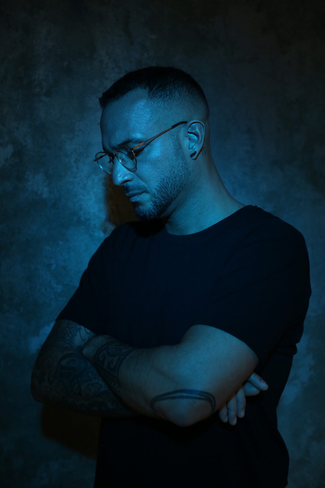 Loco Dice in round glasses and a black t-shirt, arms crossed and head lowered, lit in blue against a dark wall