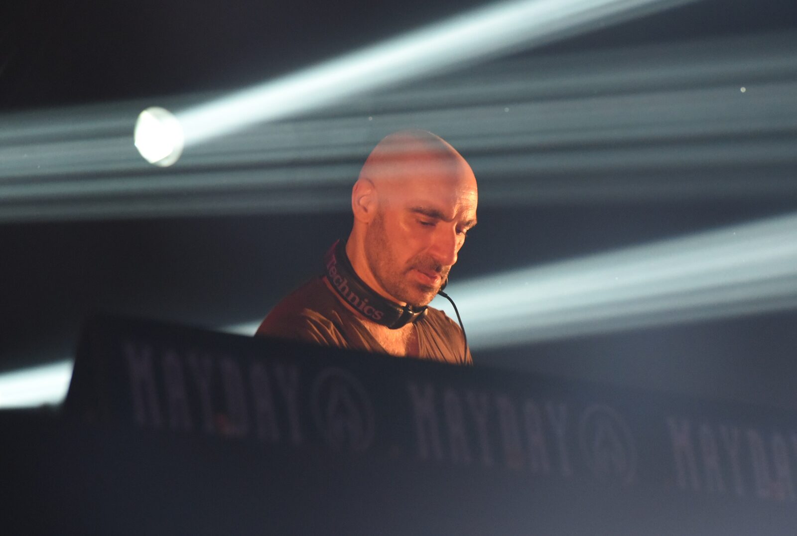 Len Faki looking down at the decks behind a Mayday-branded DJ booth, lit by white beams of light