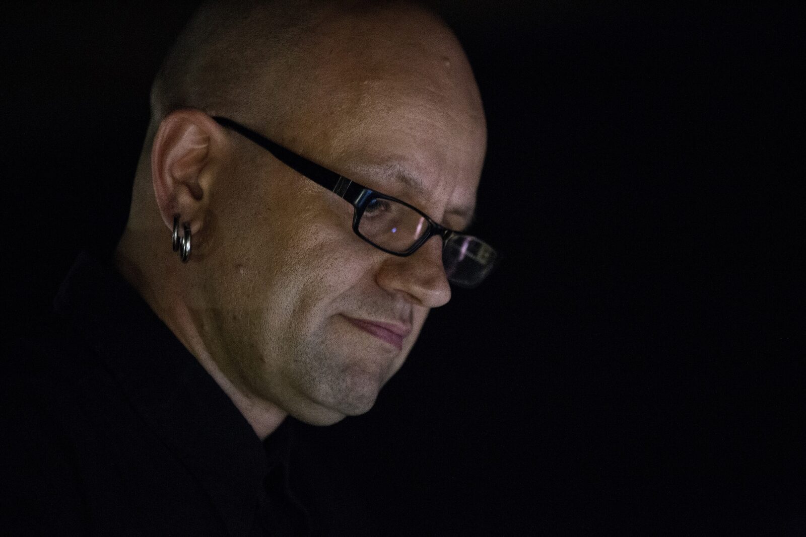 Close-up profile of a man with a shaved head, black-framed glasses and a hoop earring, lit against a black background, shown performing as Monolake in 2014