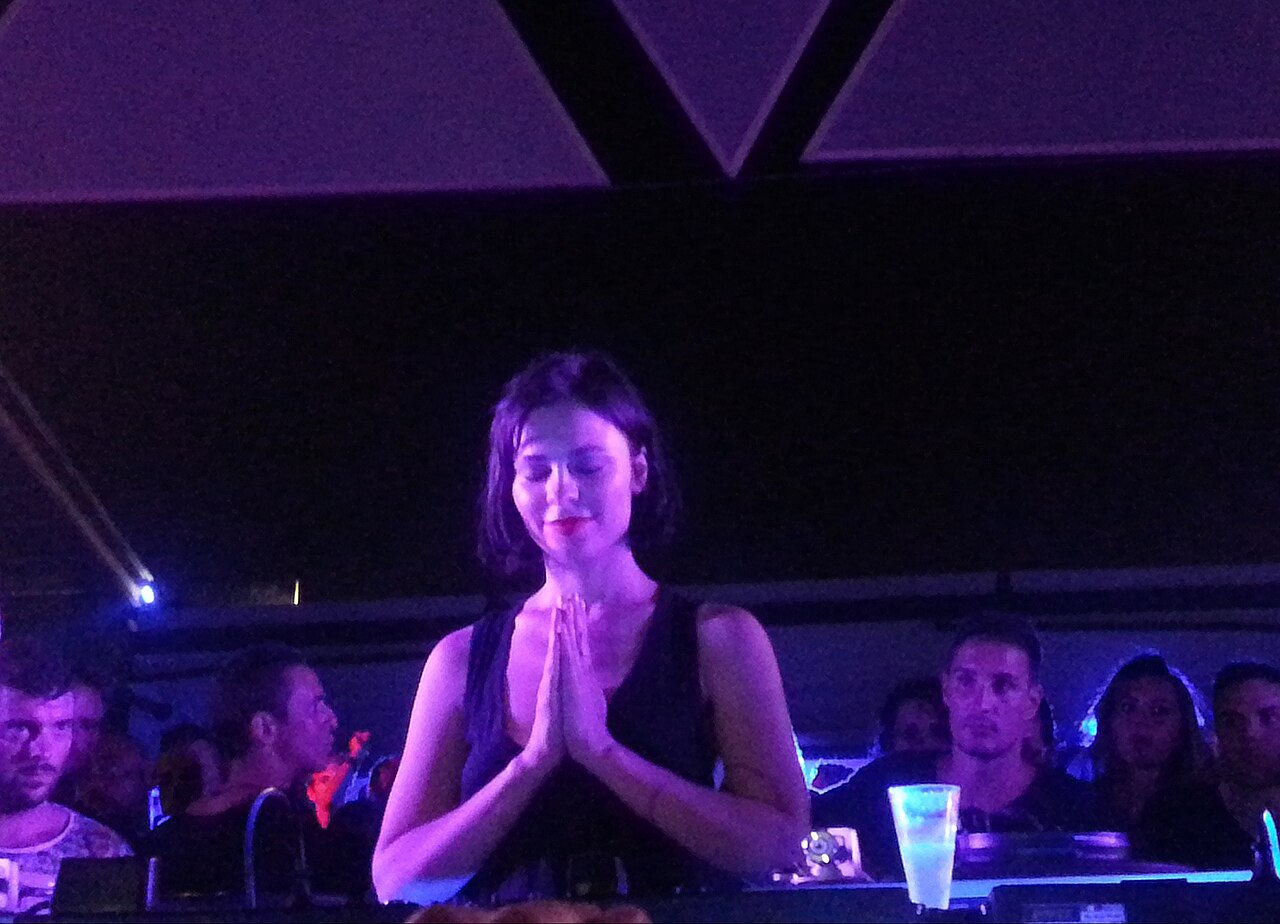 Nina Kraviz behind the decks, hands together, mid-set at a club show