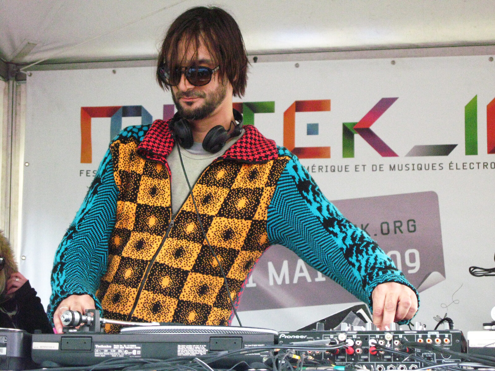Ricardo Villalobos in sunglasses and a patterned knit jacket mixing on Technics turntables in front of a 2009 festival banner