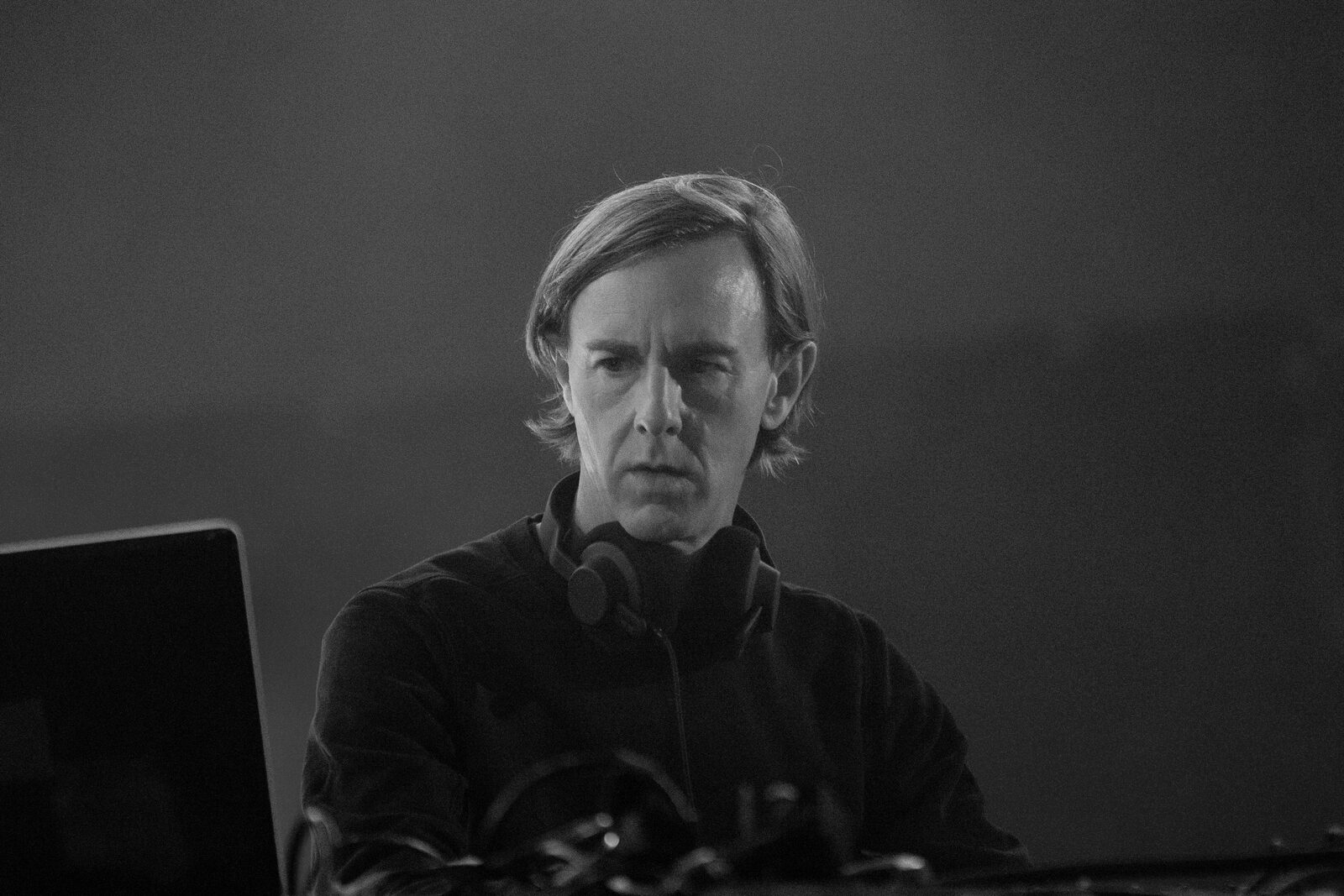 Black and white portrait of Richie Hawtin at a DJ setup with headphones around his neck, his eyes lowered toward a laptop