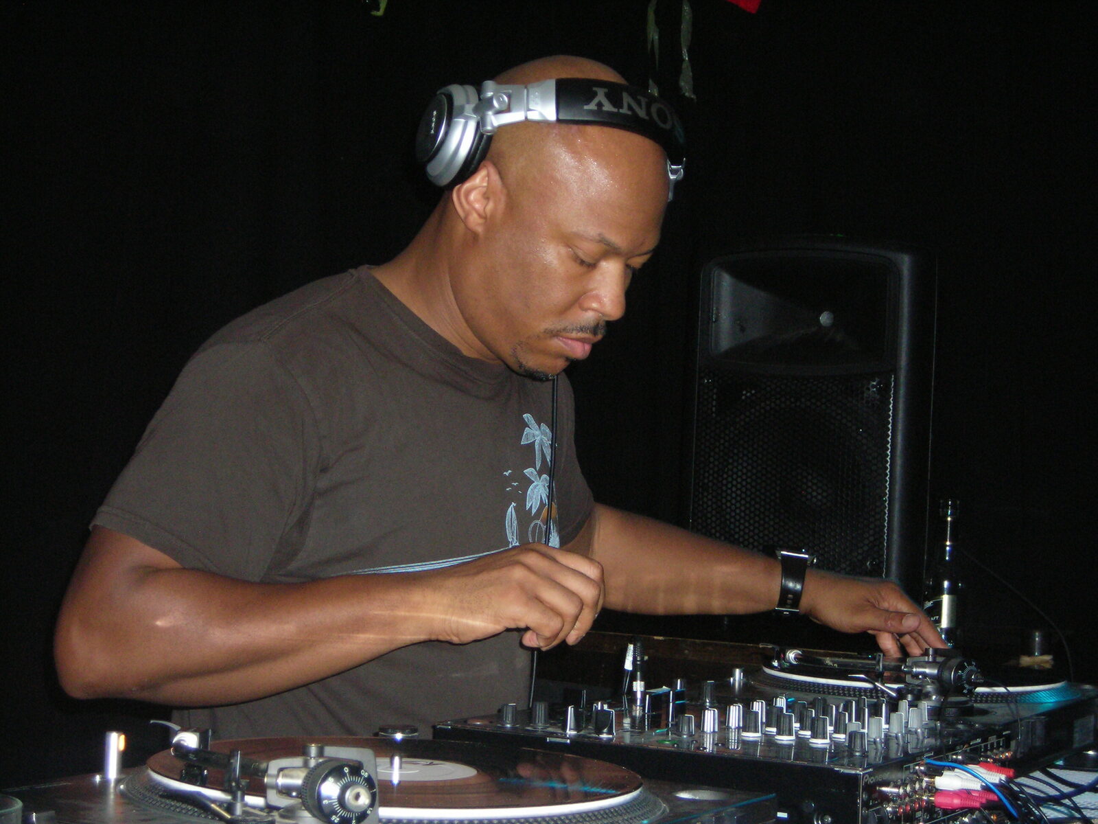 Robert Hood wearing headphones and a brown t-shirt, mixing on turntables and a DJ mixer in a dark club