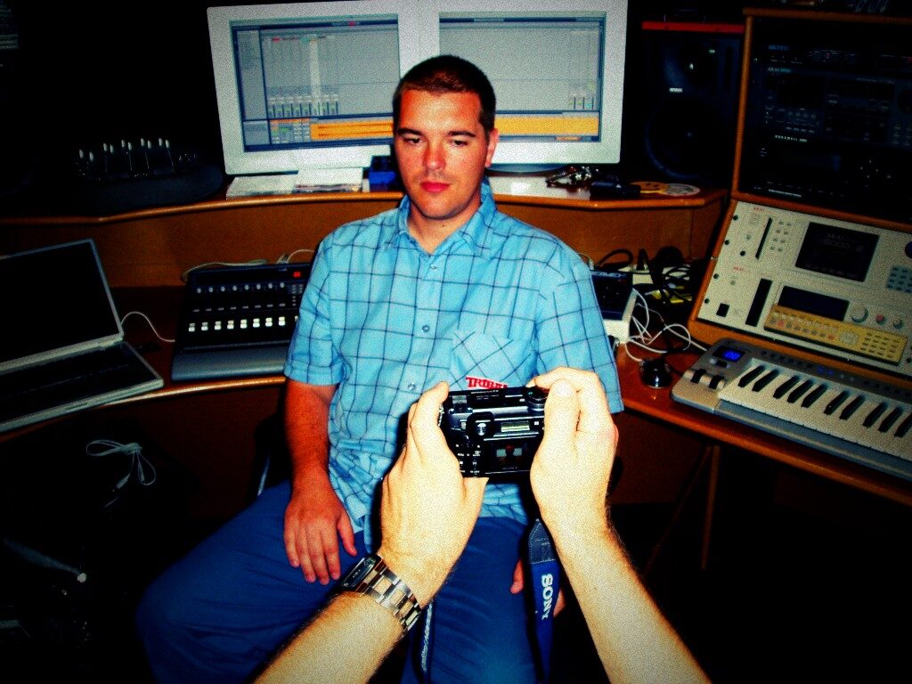 Speedy J seated in a home studio in front of two computer monitors showing music software, with a keyboard and sampler beside him and a camera held in the foreground