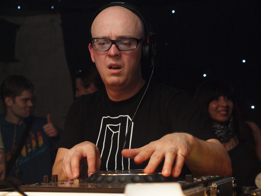 Stephan Bodzin wearing headphones and glasses, leaning over a DJ mixer in a dark club