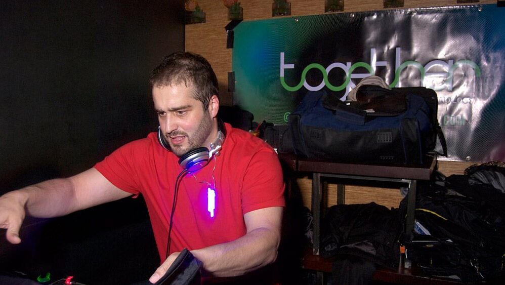 A DJ in a red T-shirt with headphones around his neck reaches toward the decks in front of a banner reading Together