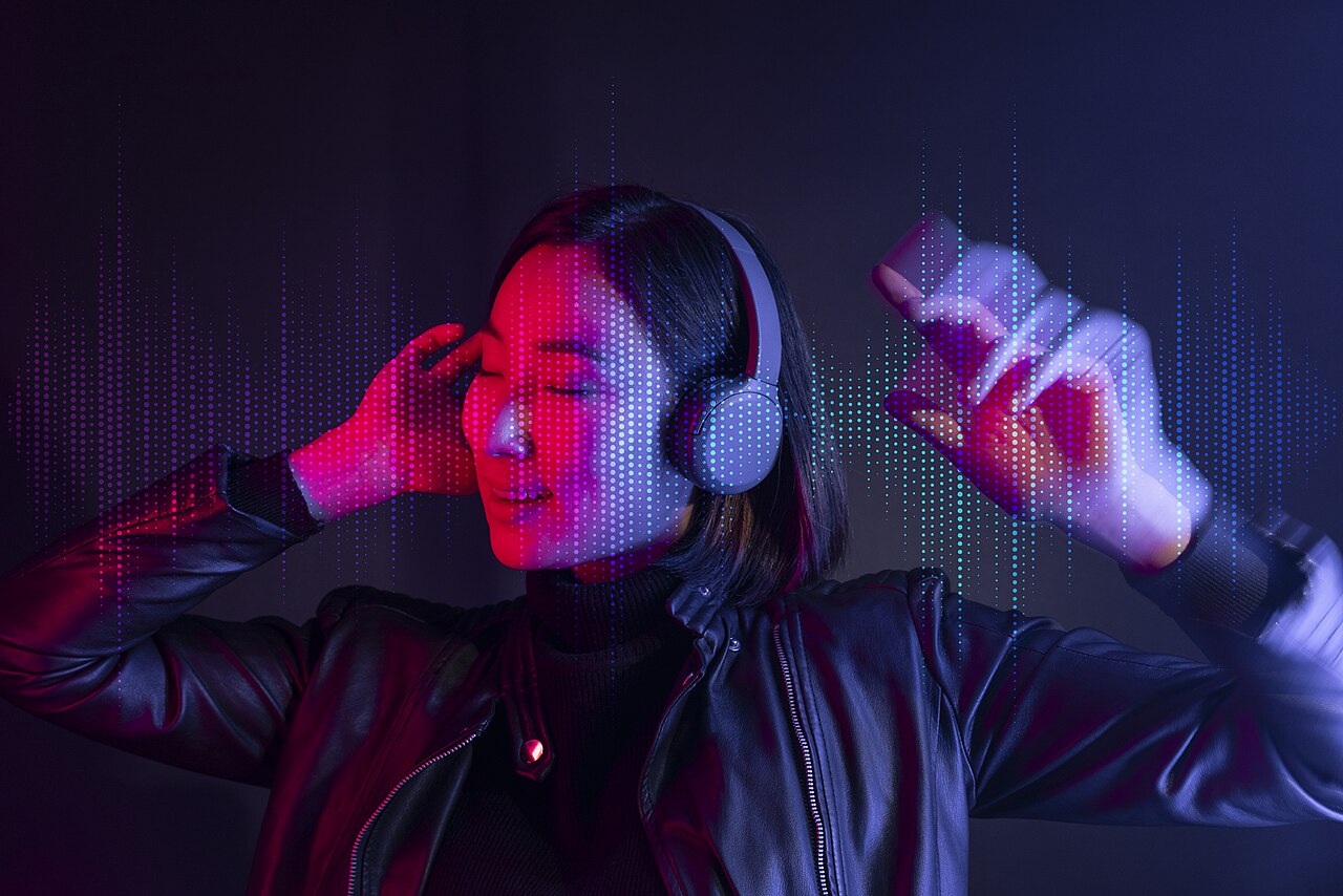 A woman listening to music through wireless headphones under neon light. Photo by Vu Hoang, CC BY 2.0, via Wikimedia Commons.