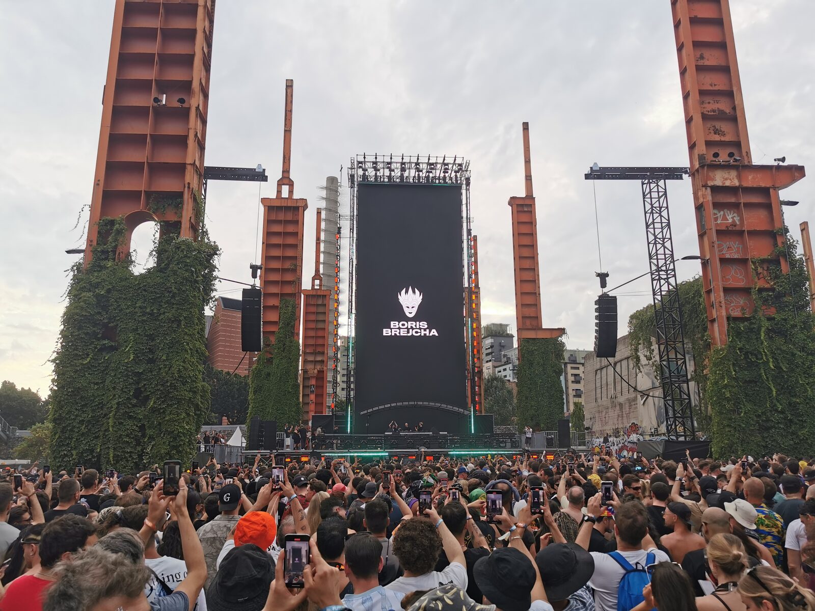 A crowd with phones raised in front of the Boris Brejcha stage at Kappa FuturFestival in Turin, with rusty steel towers covered in ivy on both sides of a tall black screen showing his joker-mask logo