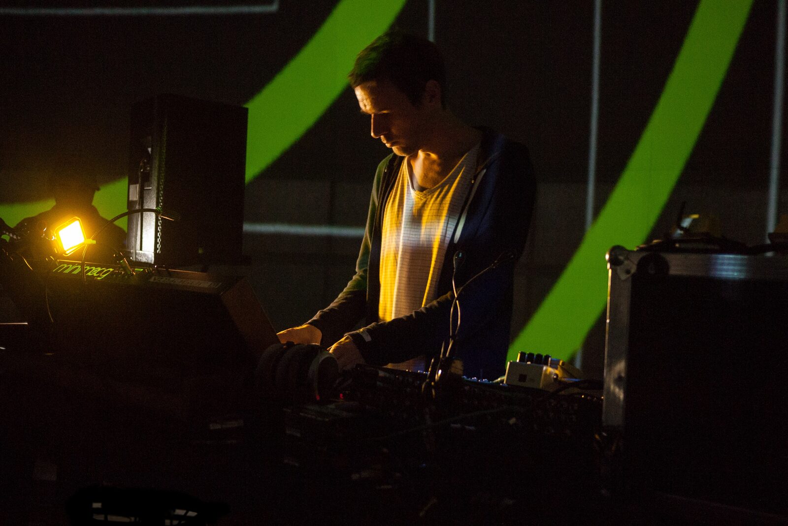 The musician Clark sound checking behind a rig of synths and mixers at MUTEK Montreal, with green projected stripes on the wall behind him.