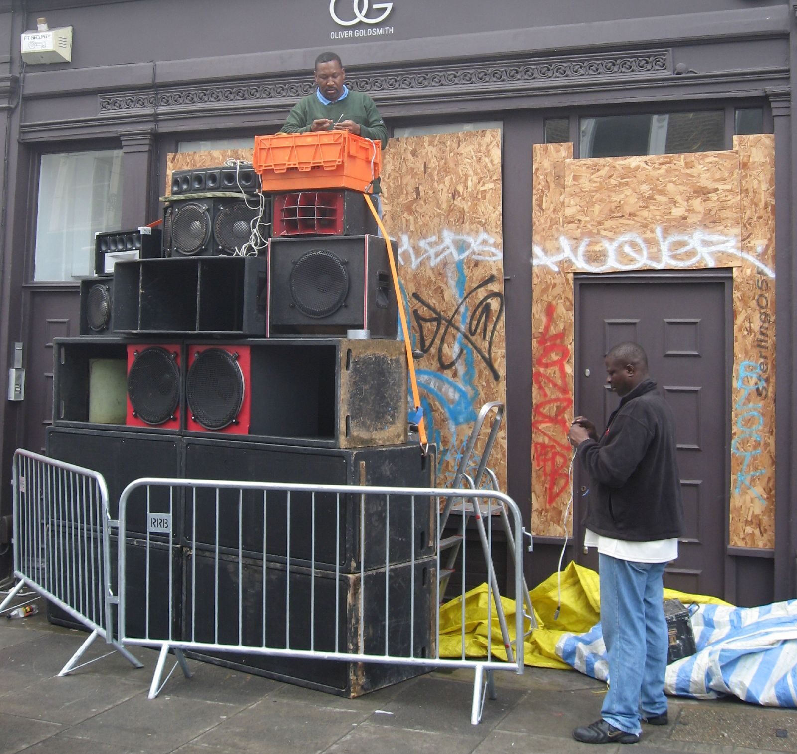 Two men setting up a tall stack of bass bins and horn speakers in front of a boarded-up shopfront