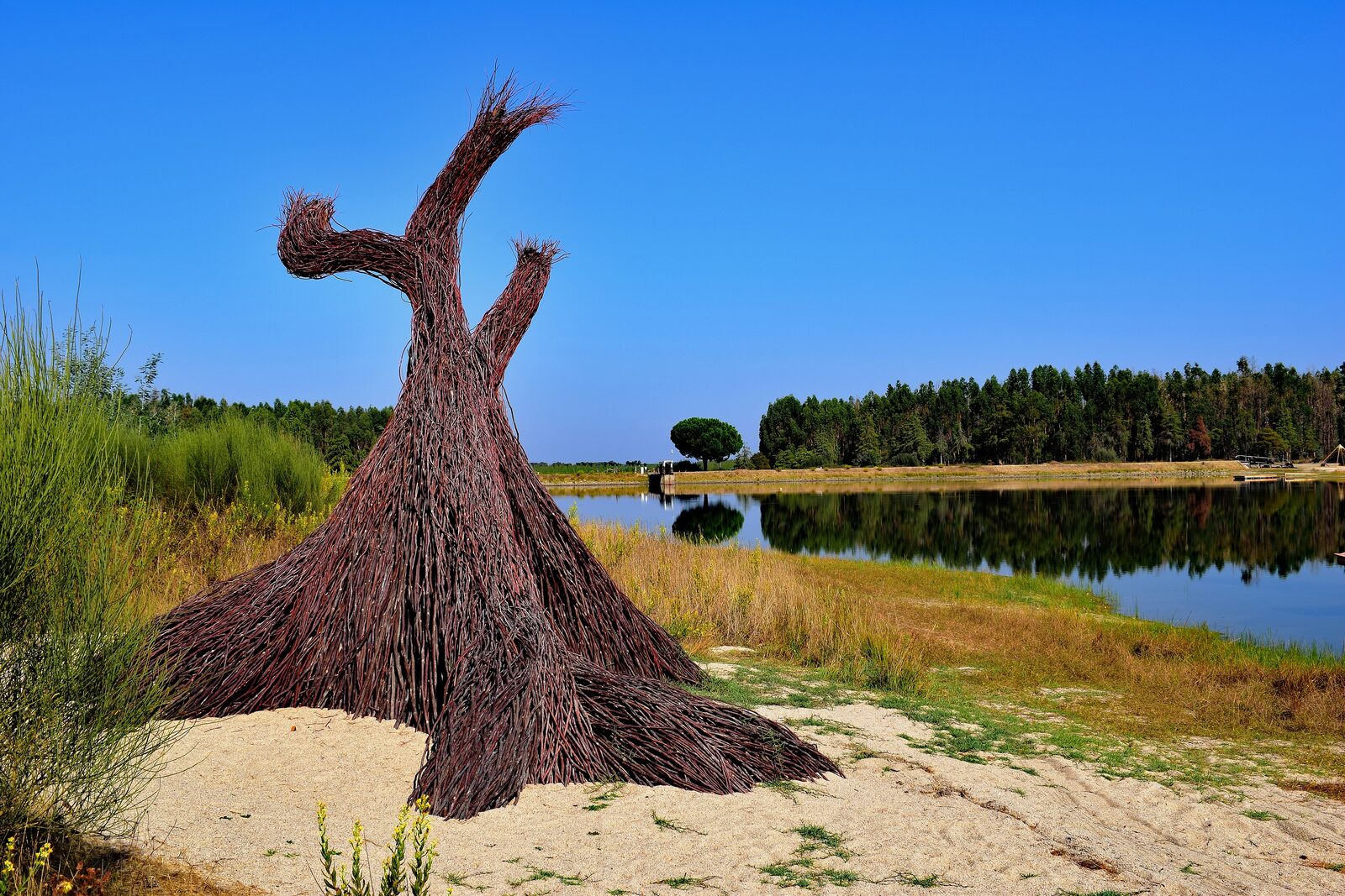 A woven willow sculpture on a sandy shore beside the water at Barragem das Nascentes in Crato, Portugal, the reservoir site of Waking Life.