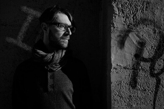 Black and white portrait of Anton Kubikov wearing glasses and a scarf, lit against a graffiti-marked wall