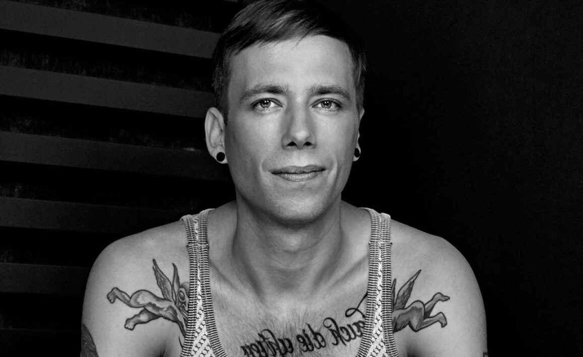 Black-and-white close-up portrait of Dirty Doering, showing his shaved-side haircut, ear gauges and chest tattoos, looking directly at the camera