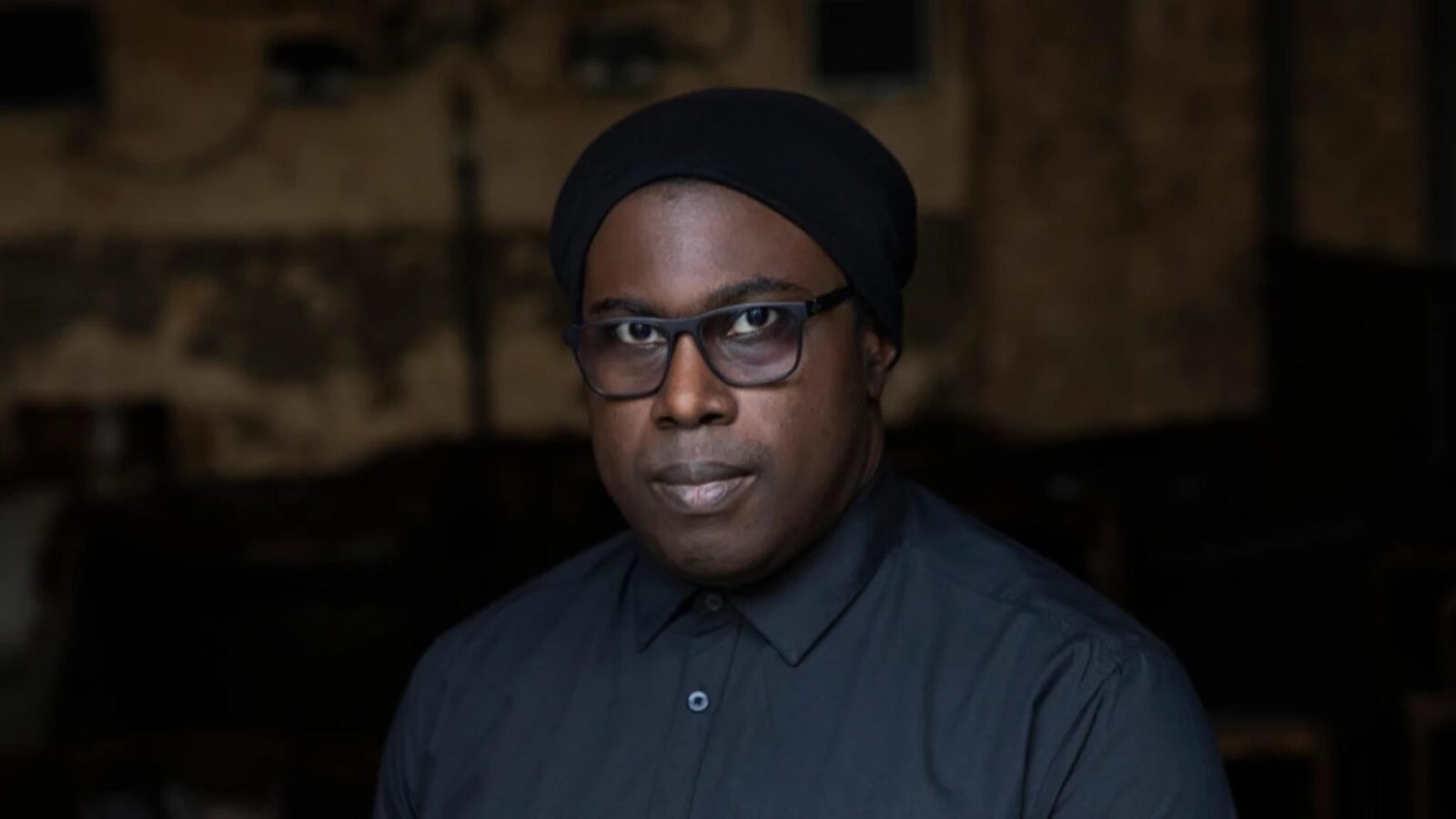 Fred P wearing a black head wrap and dark-framed glasses, photographed against a dimly lit, ornately decorated interior