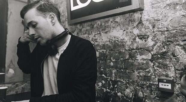 Black and white photo of James Dexter with headphones around his neck, leaning against a DJ booth in a brick-walled venue