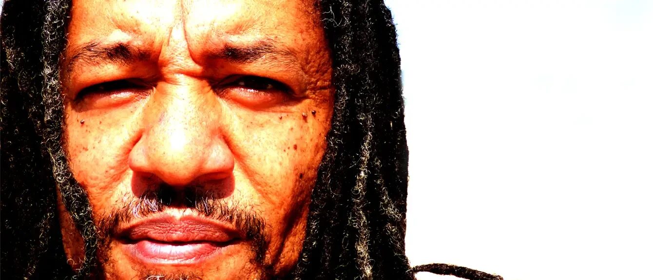 Close-up, high-contrast portrait of Jus-Ed with long dreadlocks looking directly into the camera against a bright, sunlit background