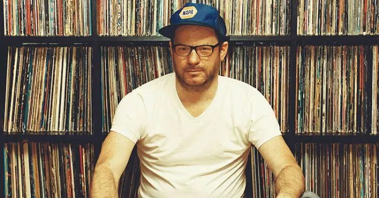 Kevin Yost wearing a blue NAPA trucker cap and glasses, seated in front of a wall of shelved vinyl records