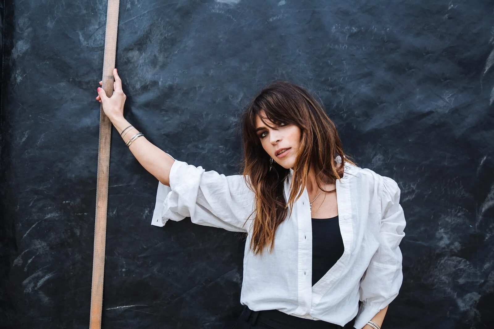 Lola Palmer looking toward the camera with one arm raised against a dark canvas backdrop, wearing an oversized white shirt over a black top