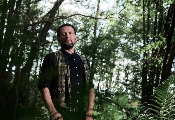 Mollono.Bass standing among trees in a forest, wearing a dark patterned vest over a black shirt