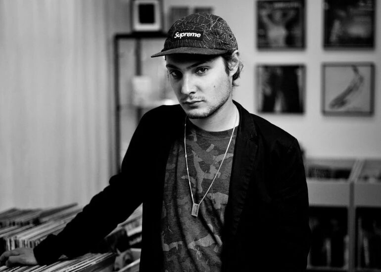 Motor City Drum Ensemble (Danilo Plessow) in a black and white photo, wearing a Supreme cap and a chain necklace, browsing vinyl records in a record store