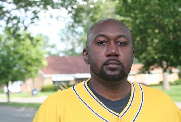 Color portrait of Rick Wade outdoors on a sunny day, wearing a yellow jersey with navy trim, standing in front of houses and trees