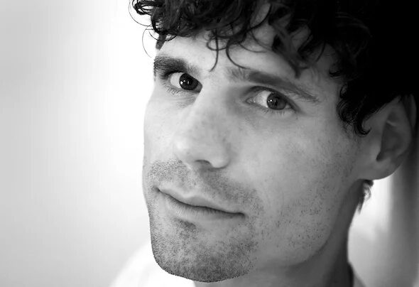 Shonky in a black and white close-up portrait, looking toward the camera with curly dark hair, against a plain light background