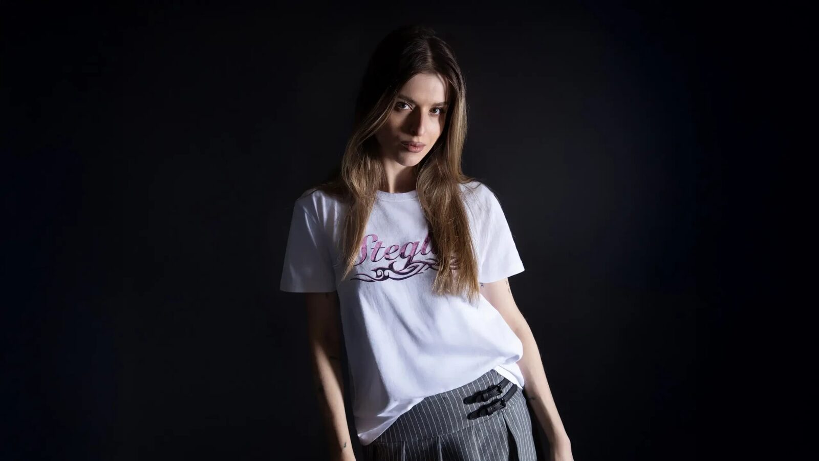 Tigerhead photographed against a black background, wearing a white T-shirt with pink script lettering and pinstriped trousers