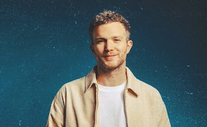 Color studio portrait of Traumer smiling gently in a tan corduroy jacket against a dark blue textured background